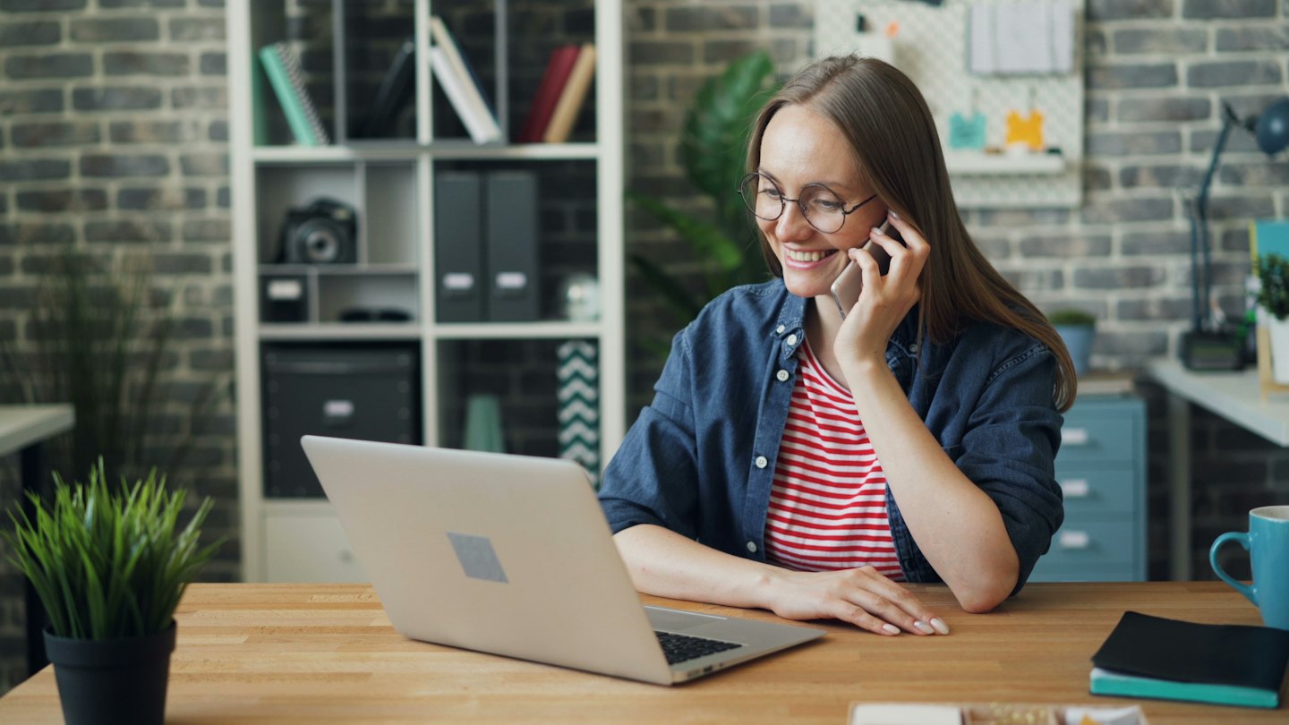 Contact Woman smiles while talking on a smartphone at her desk with a laptop.